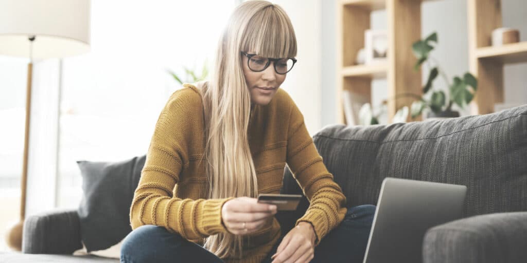 Dame med gul genser sitter i en sofa. Hun holder i et kredittkort og ser på skjermen til en laptop. Kanskje hun sjekker om hun har betalingsanmerkninger?