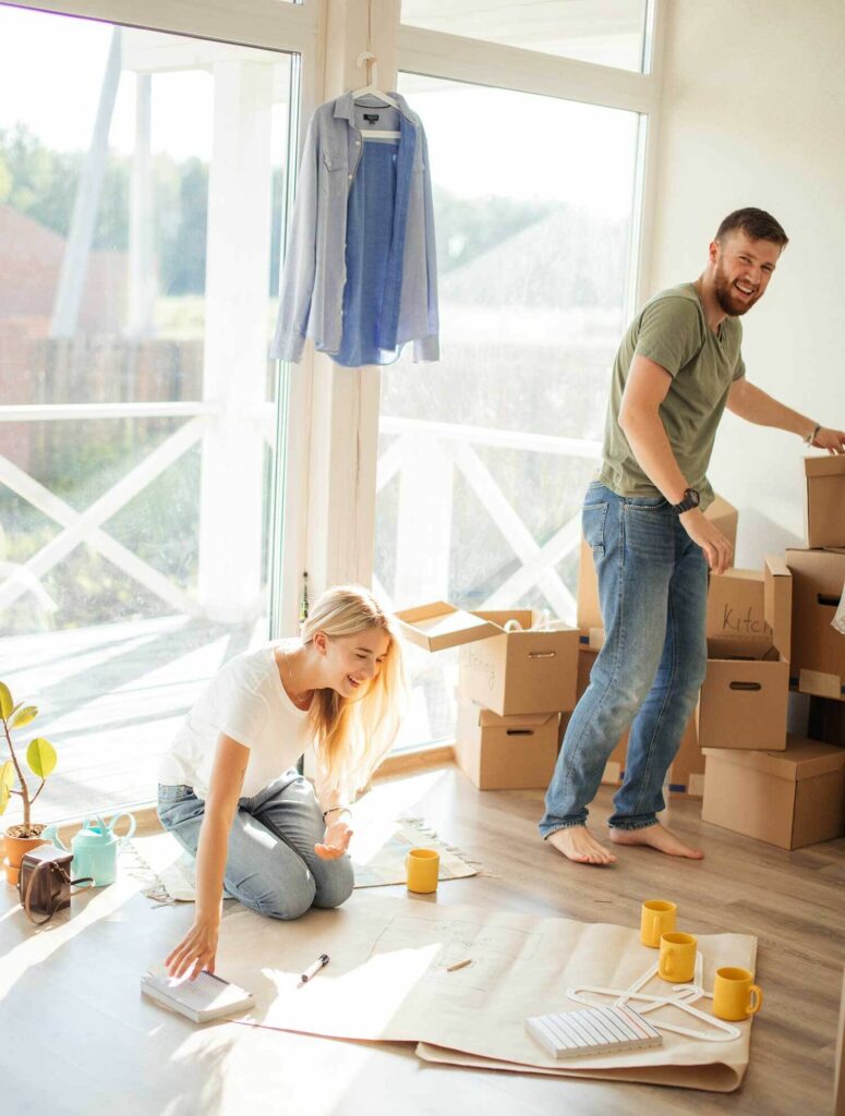 girl sits on living room floor with cups and paper and man stands upright by many cardboard boxes. young couple who have moved into a new home and are unpacking their things